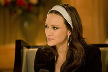 Right profile of a brown-haired teenage girl wearing a white headband, white hooped earrings and a brown coat.